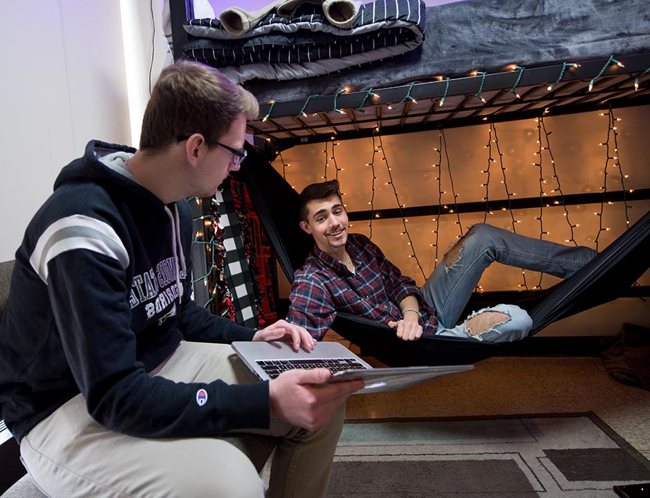 Two boys in a residence hall, one in a hammock under a loft bed and another sitting in a desk chair with a laptop.